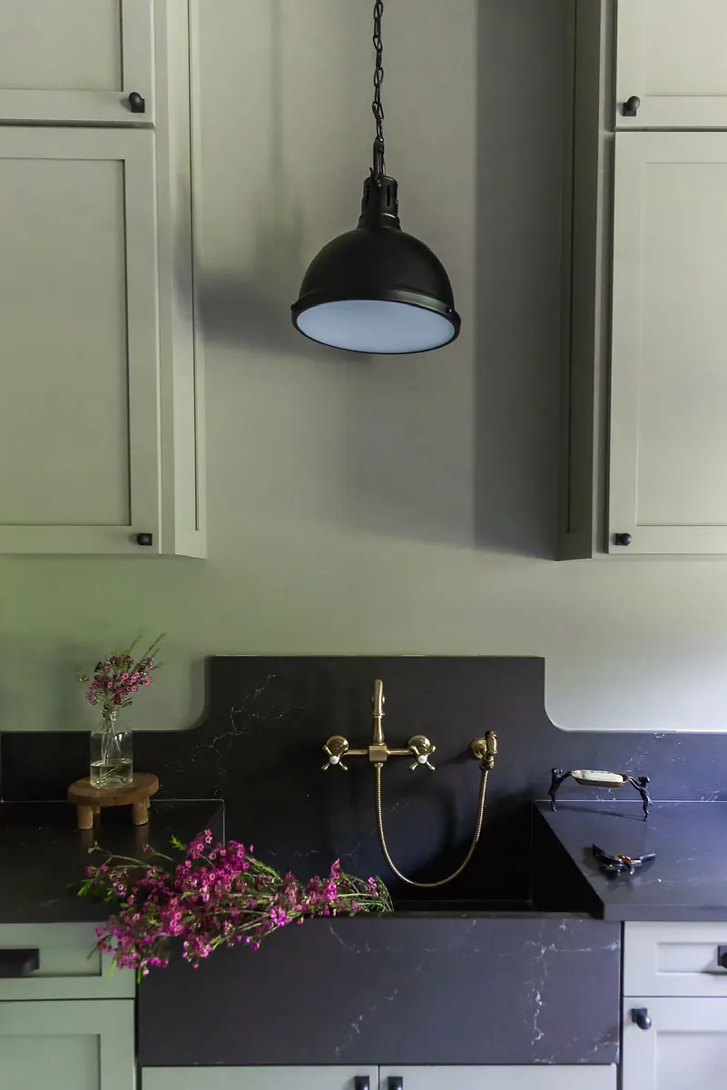 Kitchen with black countertop, grey cabinets, wall-mounted sink, brass faucet, and black pendant light
