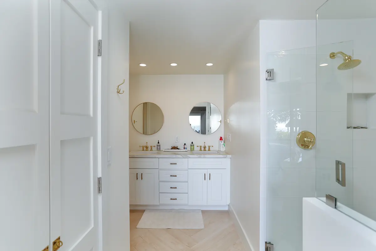 Bathroom with double vanity, round mirrors, glass shower, and white rug.