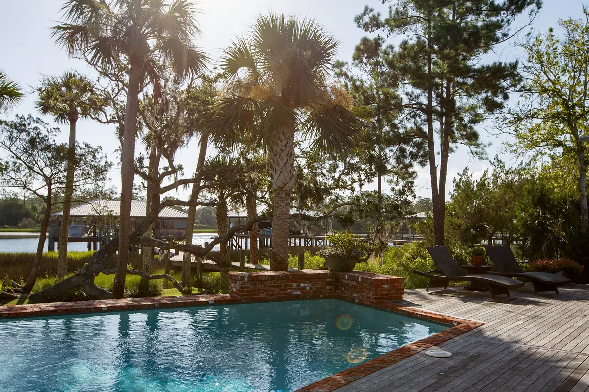 Patio with swimming pool, brick surround, wooden deck, and lounge chairs with trees and water in the background