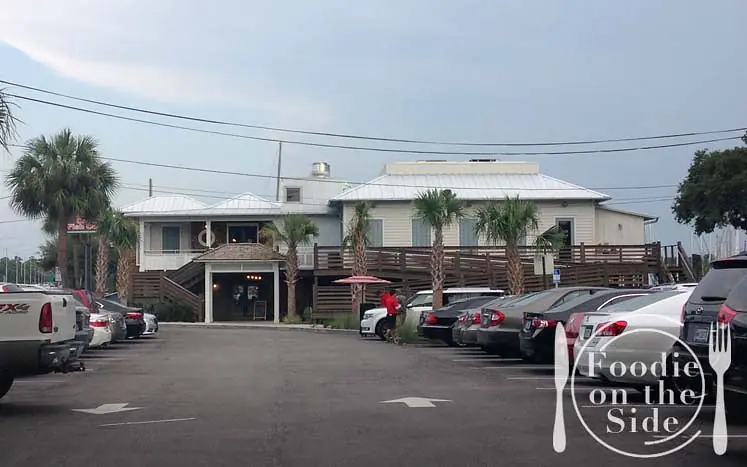 Exterior front of a restaurant with a two-story building, deck, parking lot, and palm trees.