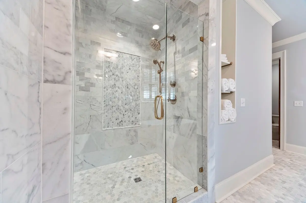 Bathroom with glass shower enclosure, marble walls, and white towels on a built-in shelf.