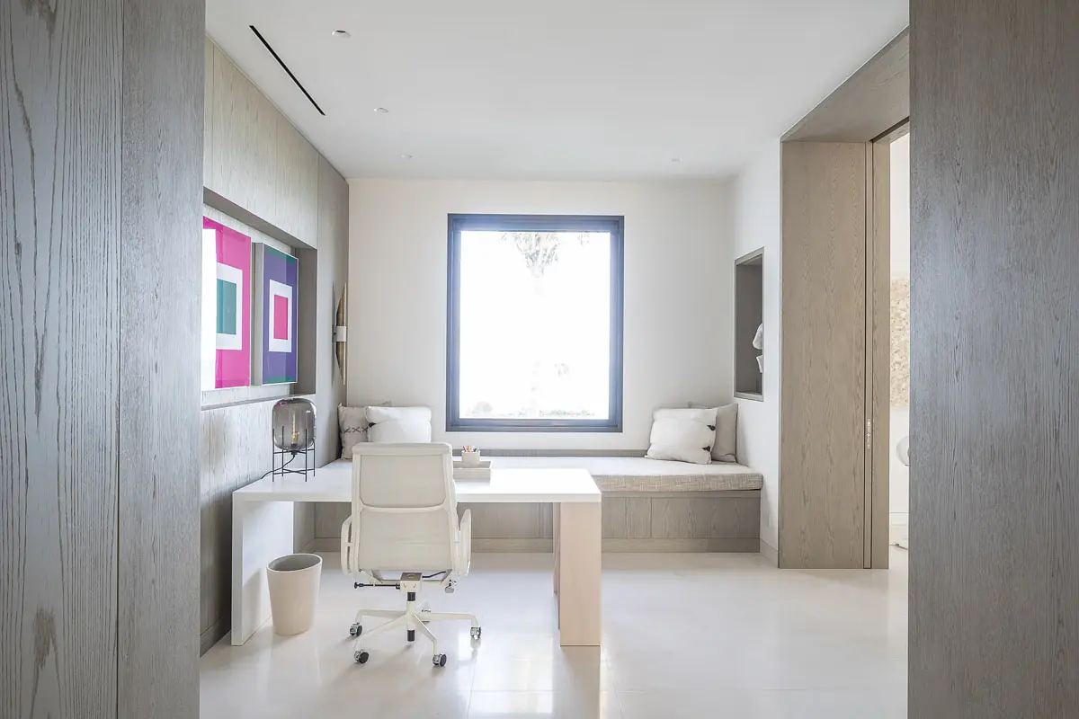 Office with white desk, ergonomic chair, built-in bench, cushions, artwork, and large window.