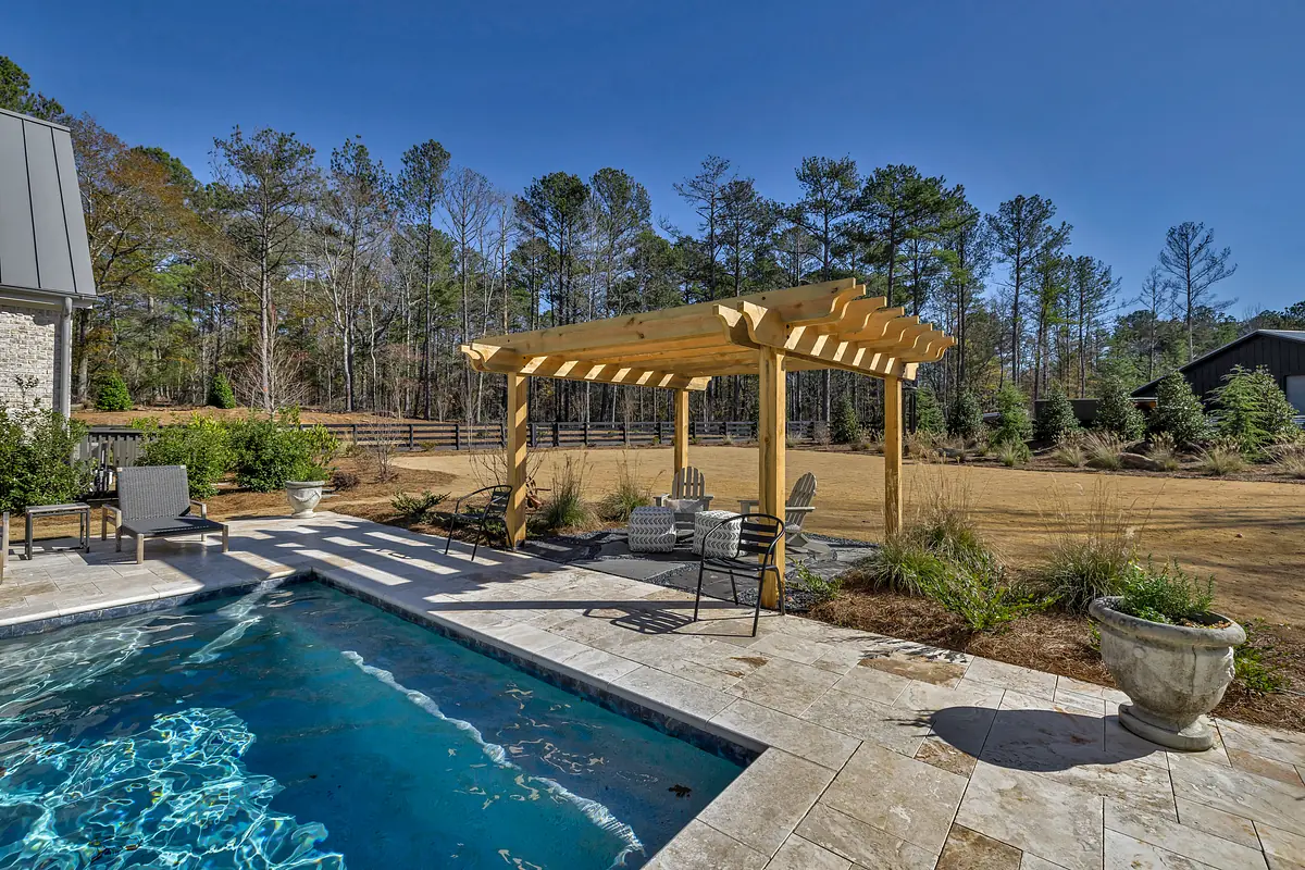 Patio with swimming pool, wooden pergola, chairs, and planters amidst greenery.