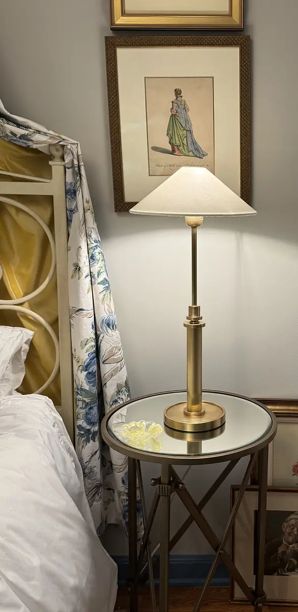 Bedroom corner with beige bed, side table, brass lamp, floral curtain, and wall artwork.