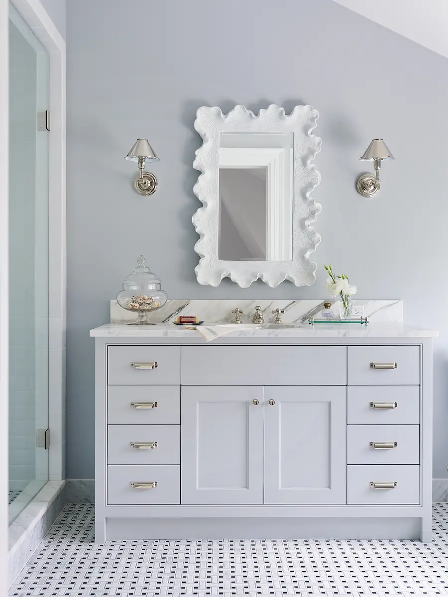 Bathroom with gray vanity, white countertop, scalloped mirror, sconces, glass shower, and black and white floor.