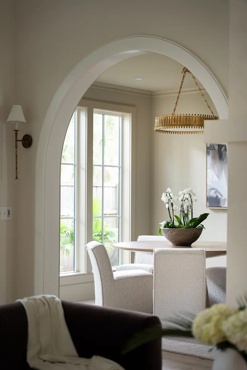 Dining room with round wooden table, upholstered chairs, chandelier, and orchids in a decorative planter.