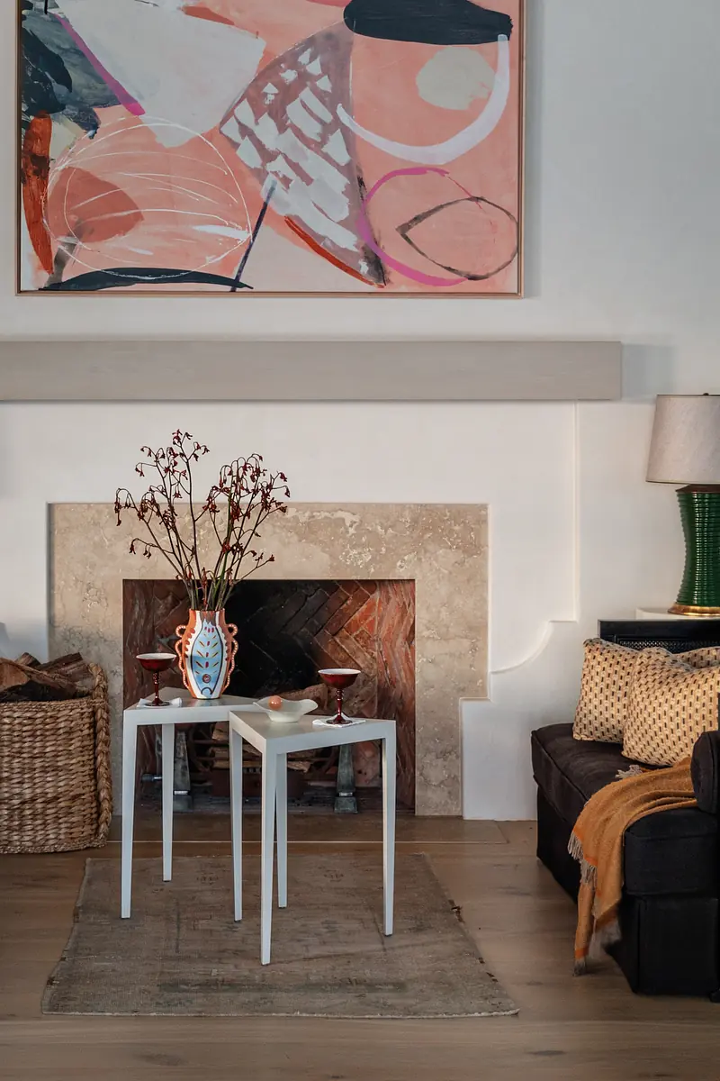 Living room with brown sofa, white side tables, decorative vase, painting, and brick fireplace.