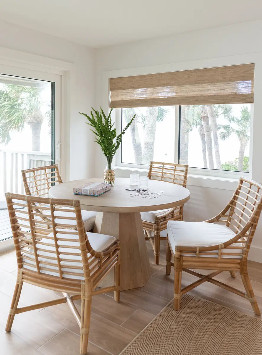 Dining room with round table, rattan chairs, vase with greenery, large windows, and natural fiber rug.