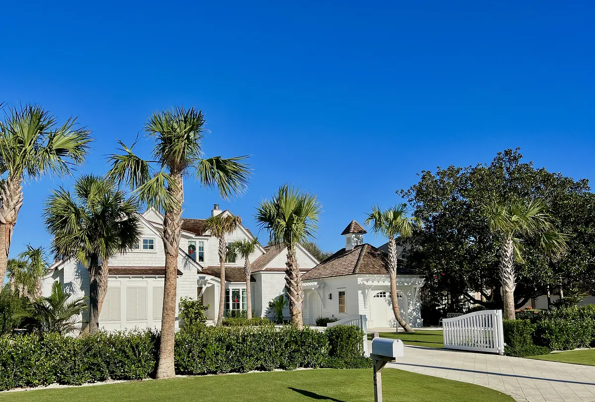 Exterior front of a house with white facade, brown roof, palm trees, and a picket fence
