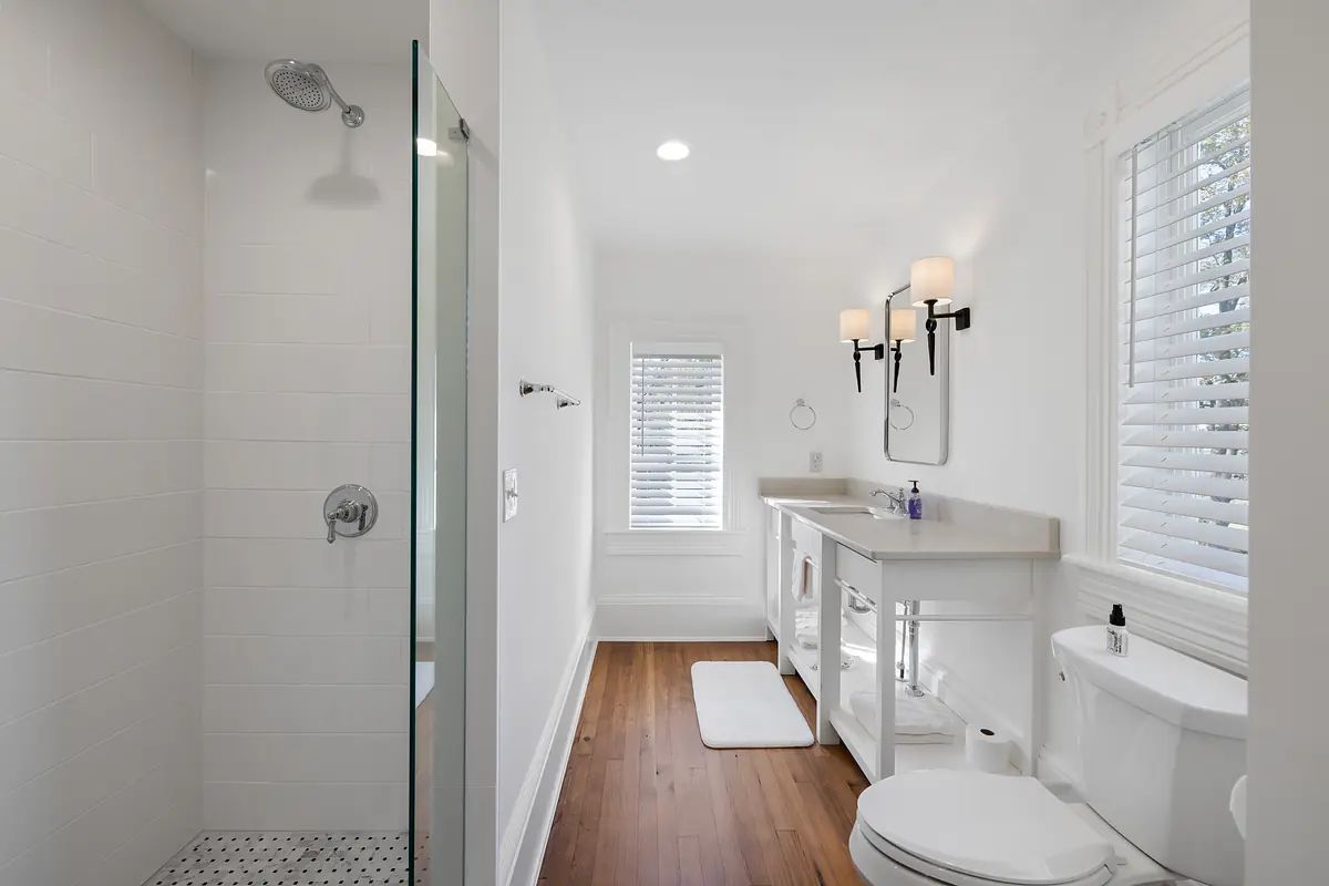 Bathroom with glass shower, white vanity, mirrors, wood floor, and window with blinds.