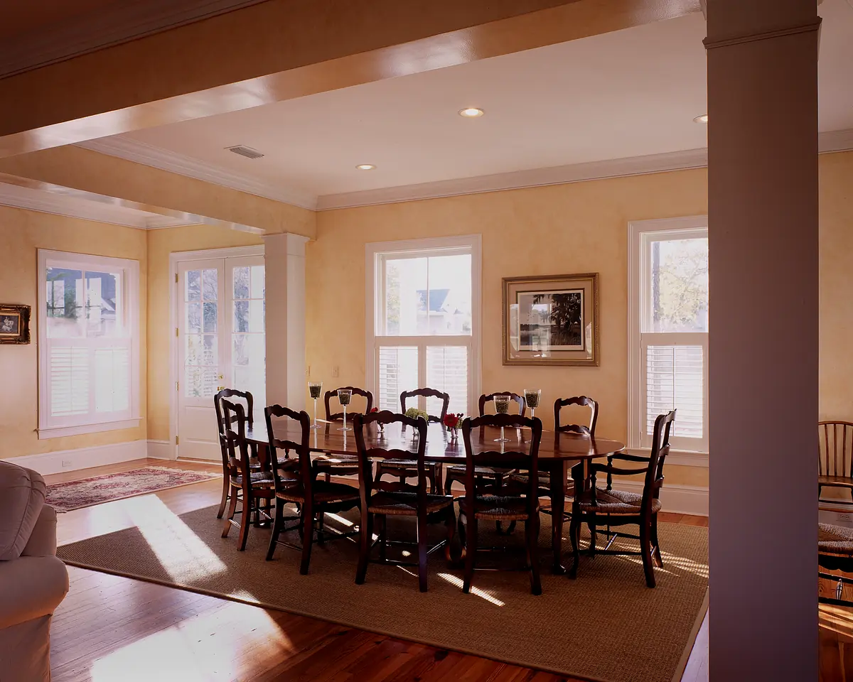 Dining room with large wooden table, twelve chairs, yellow walls, windows with shutters, and woven area rug.