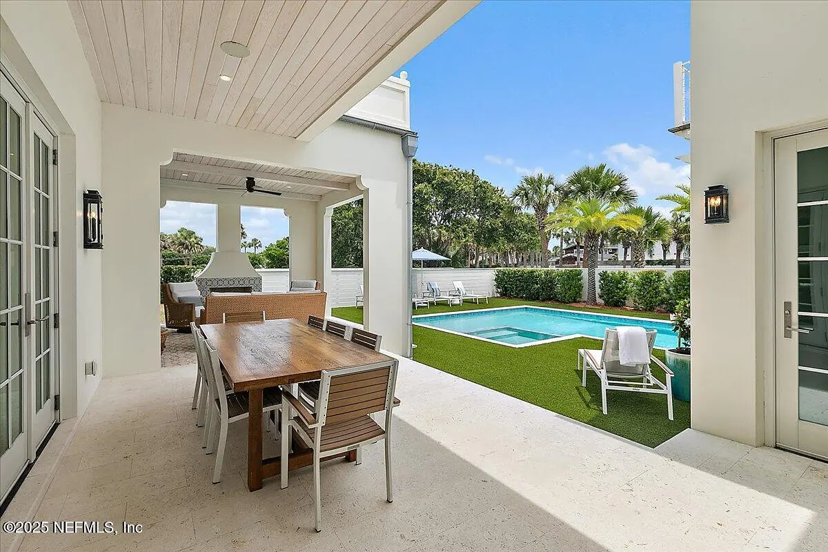 Patio with wooden dining table, chairs, pool, fireplace, and surrounding greenery.