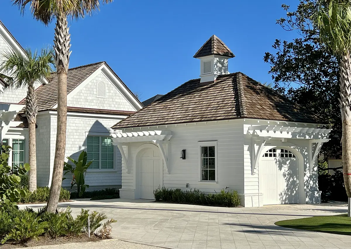 Garage with white siding, double door, cupola, palm trees, and stone tile driveway.