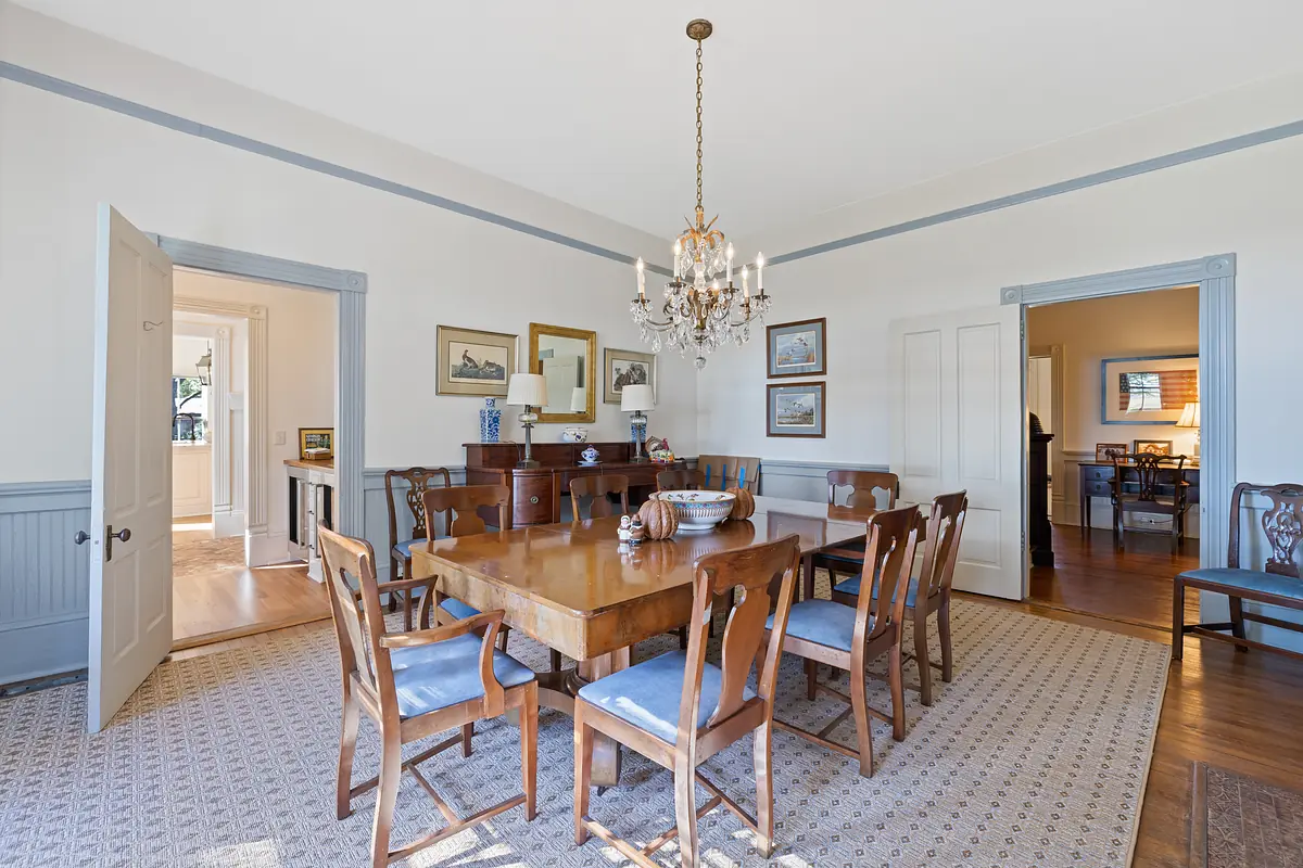 Dining room with wooden table, chairs, chandelier, artworks, sideboard, and area rug.
