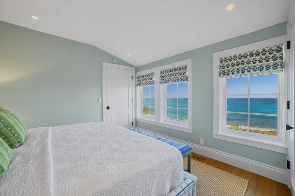 Bedroom with white quilted bed, striped cushions, blue bench, large windows, and light blue walls