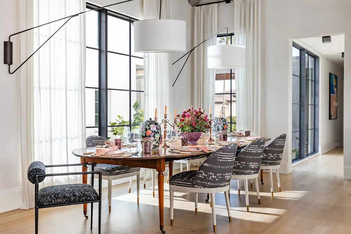 Dining room with a round table set for eight, patterned chairs, flowers, large windows, and a floor length mirror