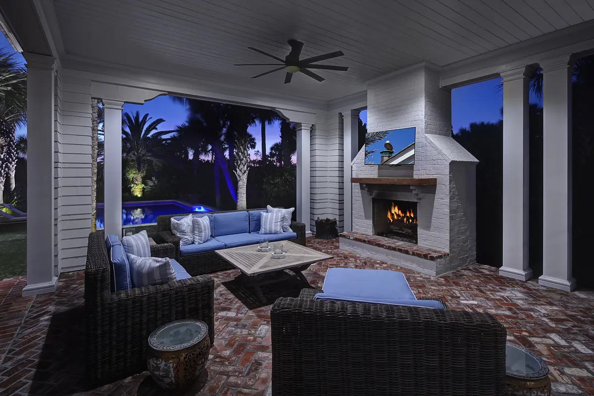 Patio with blue cushioned seating, coffee table, fireplace, and mounted television surrounded by greenery.