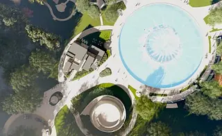 Aerial view of a park with water feature, paths, trees, and adjacent building.
