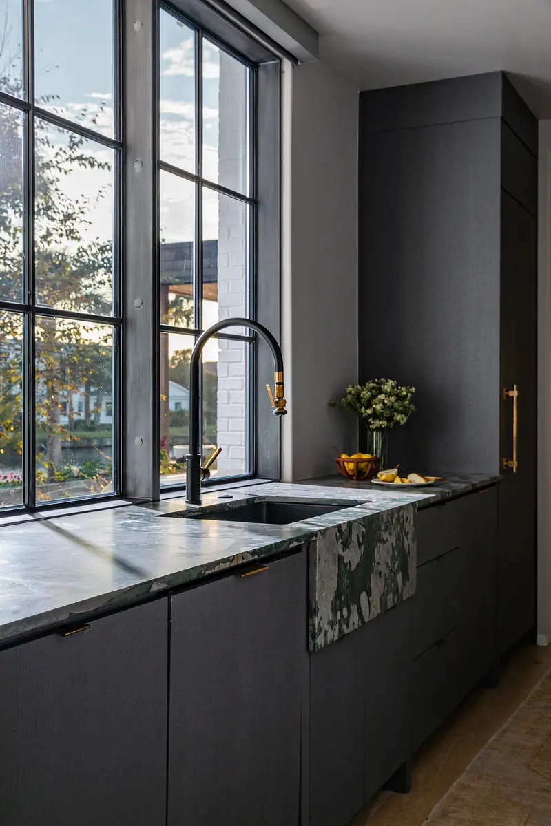 Kitchen with dark cabinetry, green marble countertop, black faucet, large window, flowers, and fruit bowl.