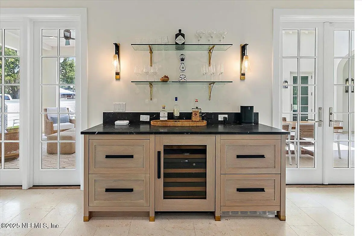 Butler's pantry with dark countertop, light wood cabinetry, wine cooler, glassware, and wall-mounted shelves.