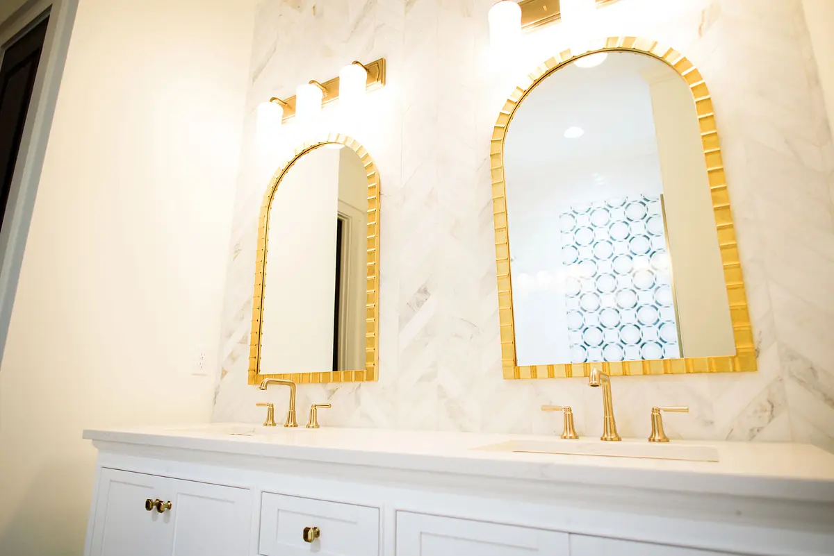 Bathroom with double vanity, arched mirrors, gold faucets, and marble tiled wall.
