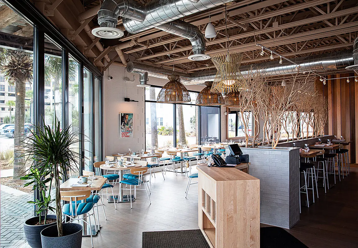 Dining room with round tables, blue chairs, bar area, large windows, exposed beams, and decorative branches.