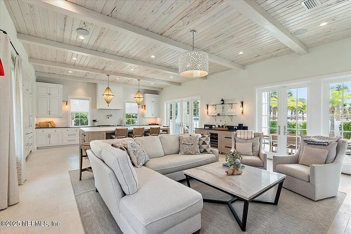 Living room with sectional sofa, coffee table, armchairs, wood beams, large windows, and kitchen view