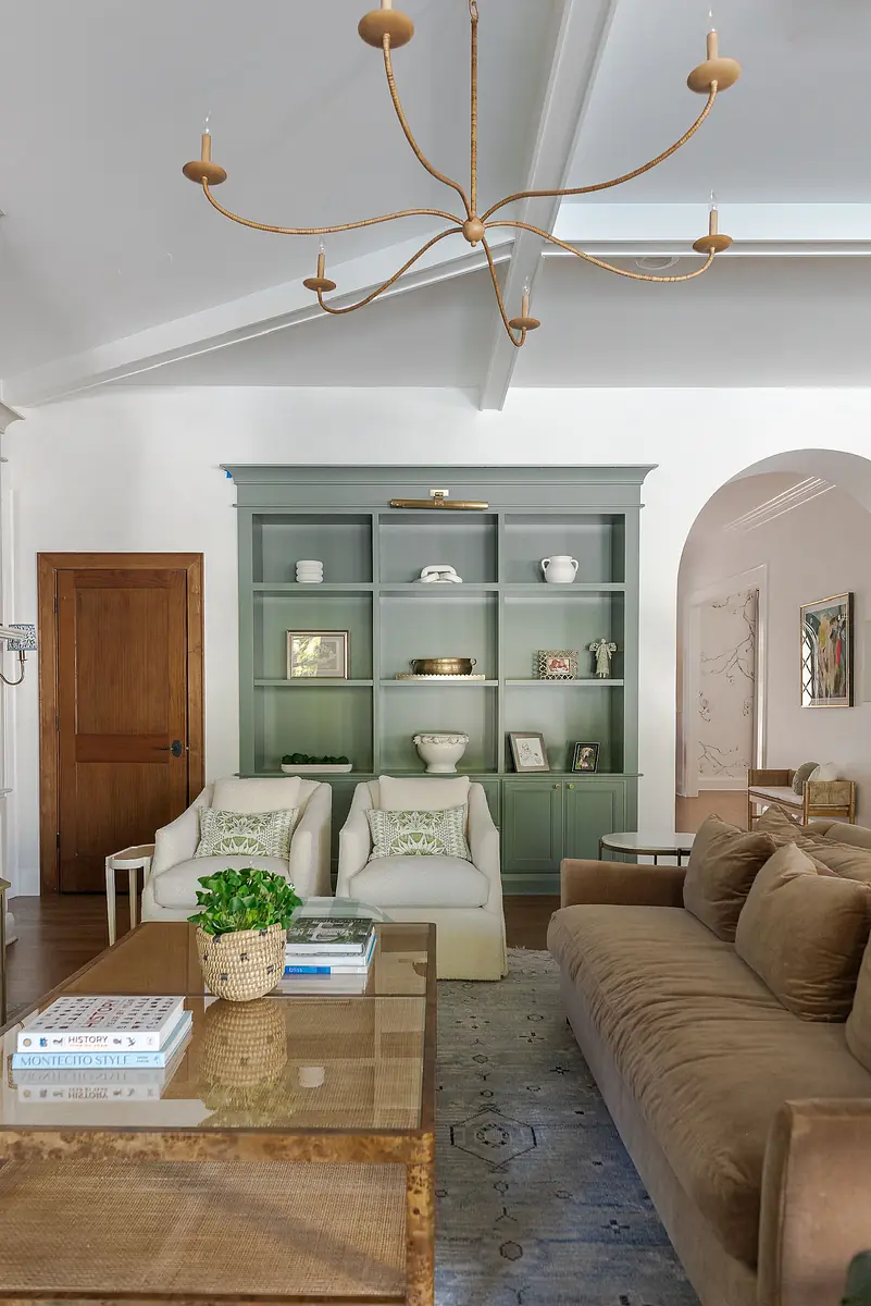 Living room with brown sofa, glass coffee table, green shelves, armchairs, and artwork on white walls.