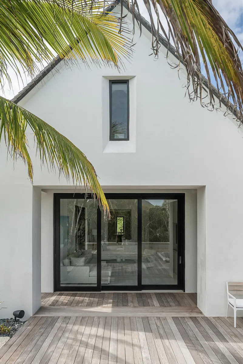 Exterior front view with white facade, black framed sliding doors, wooden decking, and palm trees.