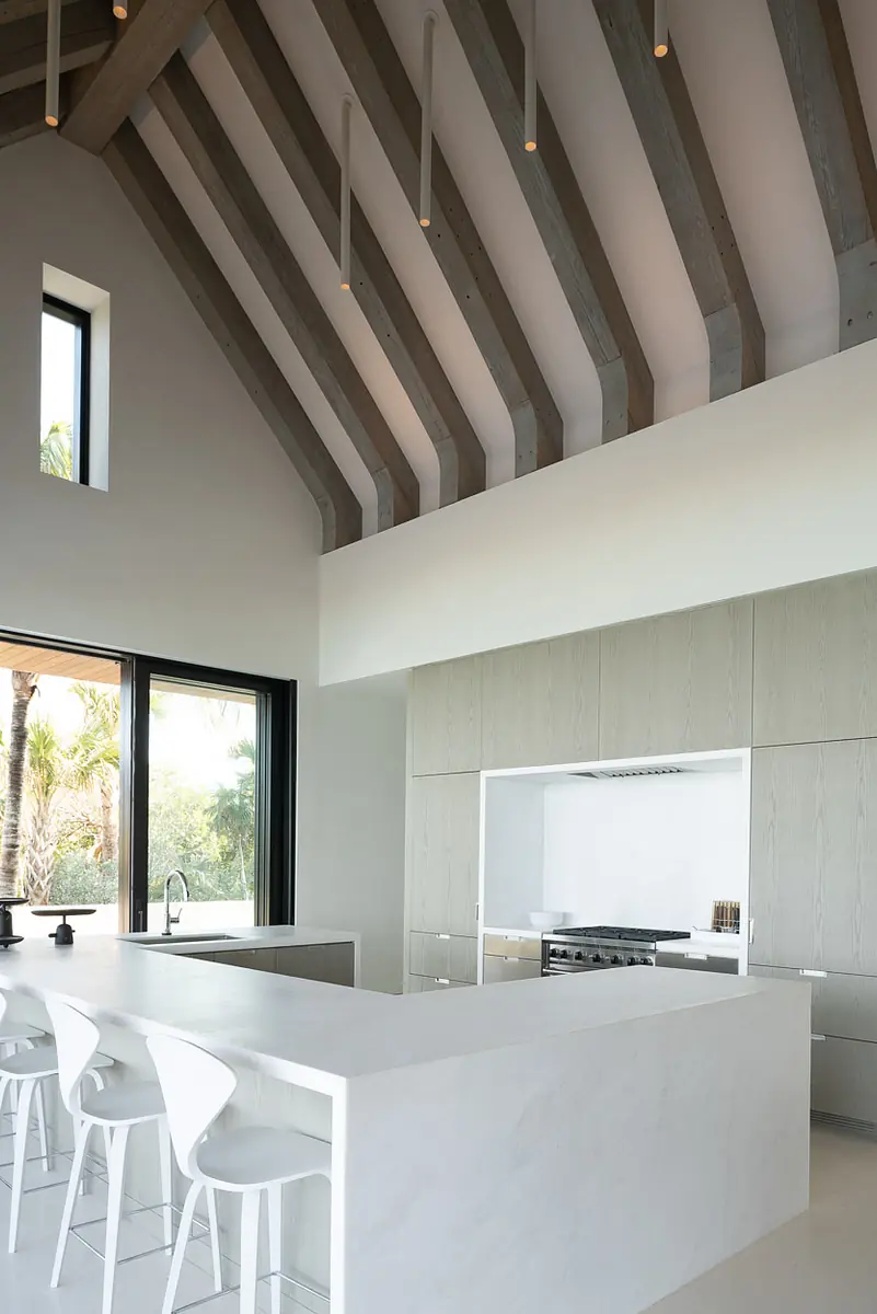 Kitchen with white countertop island, bar stools, high wooden beams, and large windows.