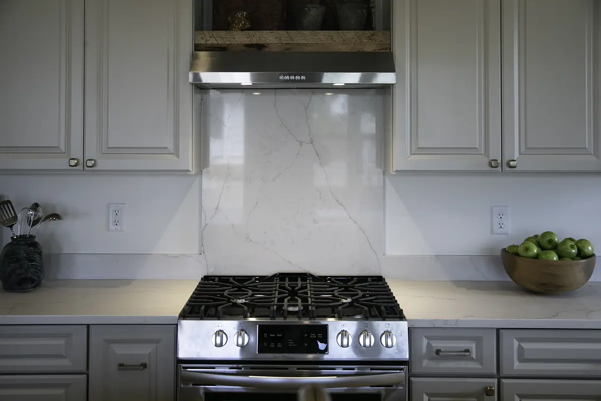 Kitchen with gray cabinetry, white countertops, gas stove, marble backsplash, and wooden bowl of green apples.