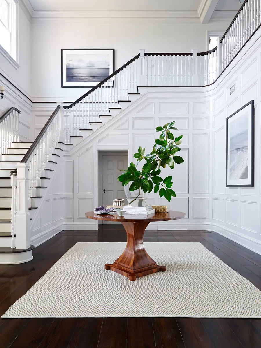 Foyer with round wooden table, potted plant, area rug, wainscoted walls, and a staircase.
