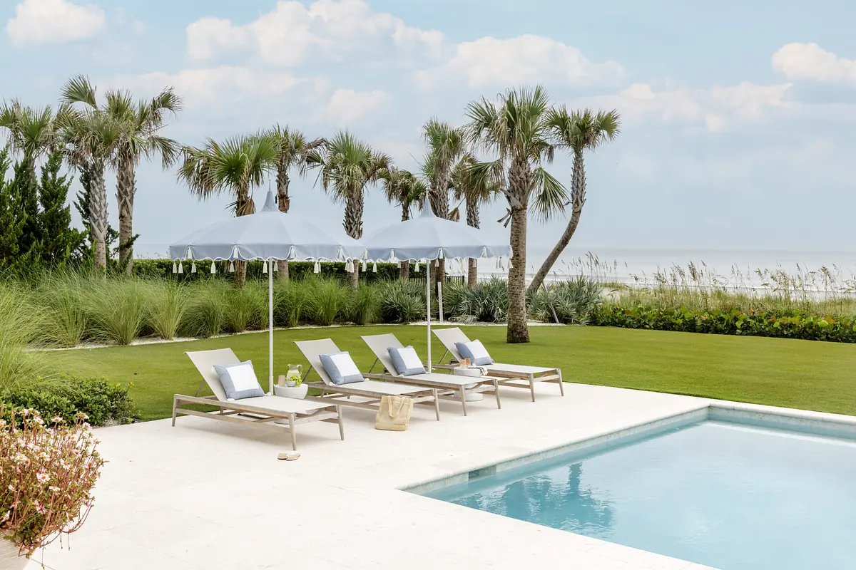 Patio with four gray lounge chairs, blue umbrellas, swimming pool, palm trees, and grassy landscaping.