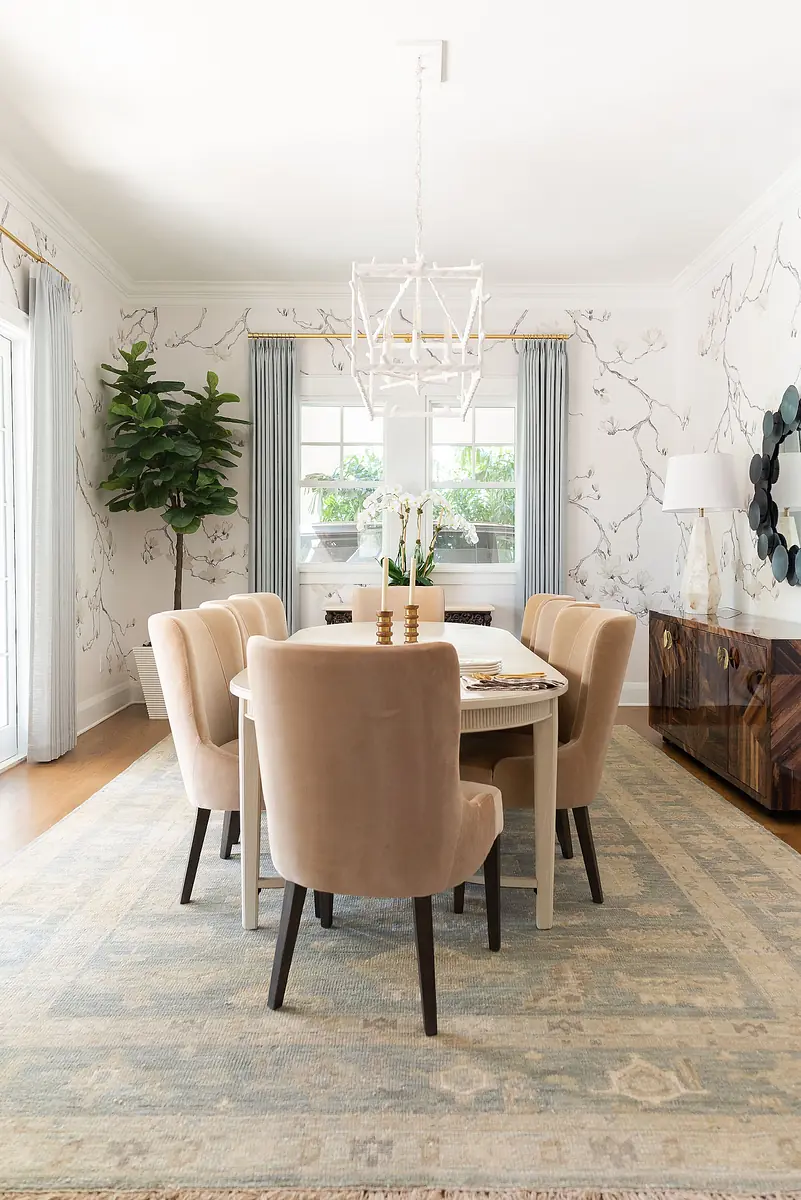 Dining room with circular table, six chairs, gold candlesticks, large rug, curtains, plant, and wall-mounted mirror