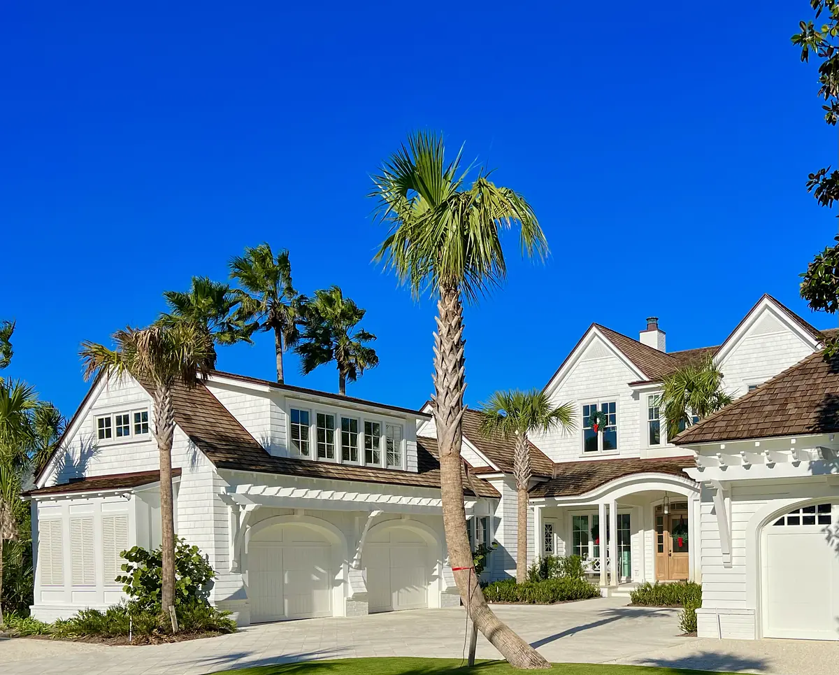 Exterior front view of a house with white siding, brown roof, large windows, driveway, and palm trees.