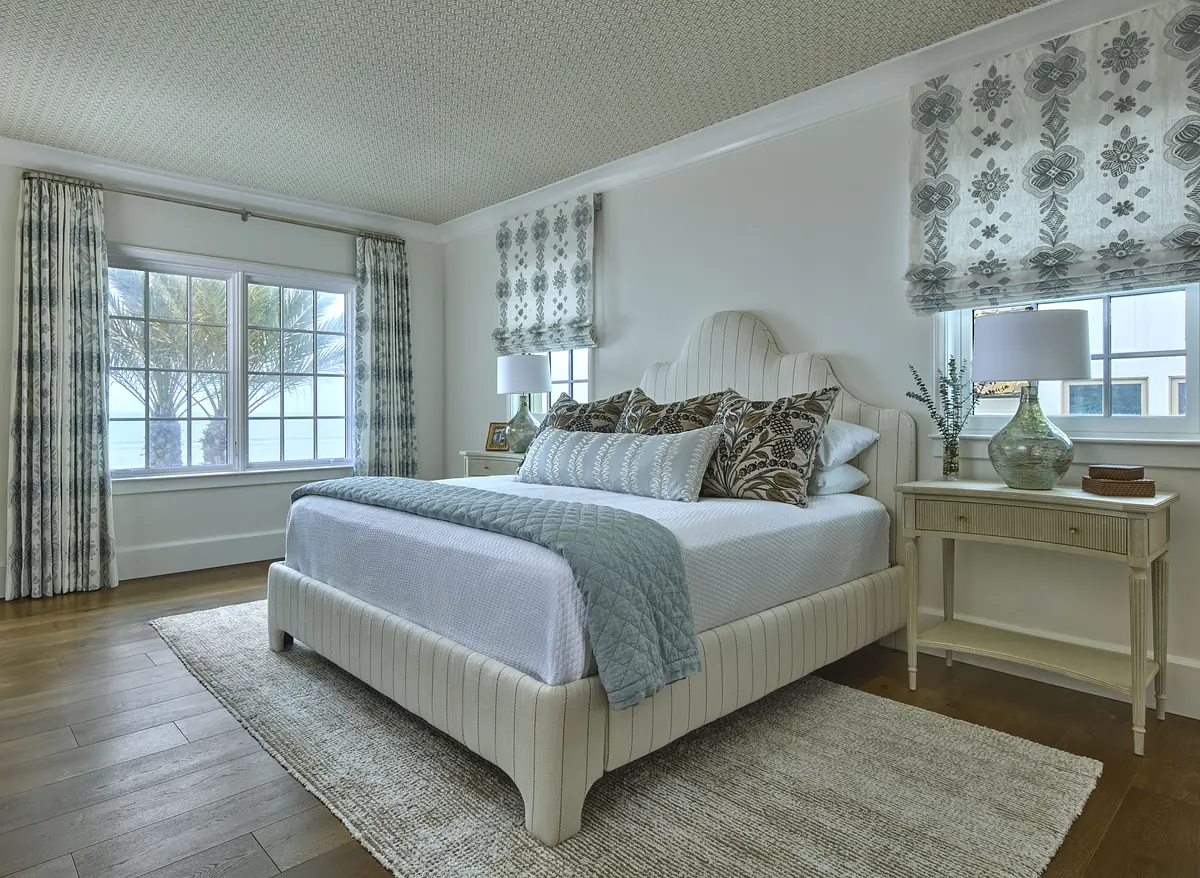 Bedroom with bed, blue quilt, nightstands, lamps, large windows, and patterned rug on hardwood floor