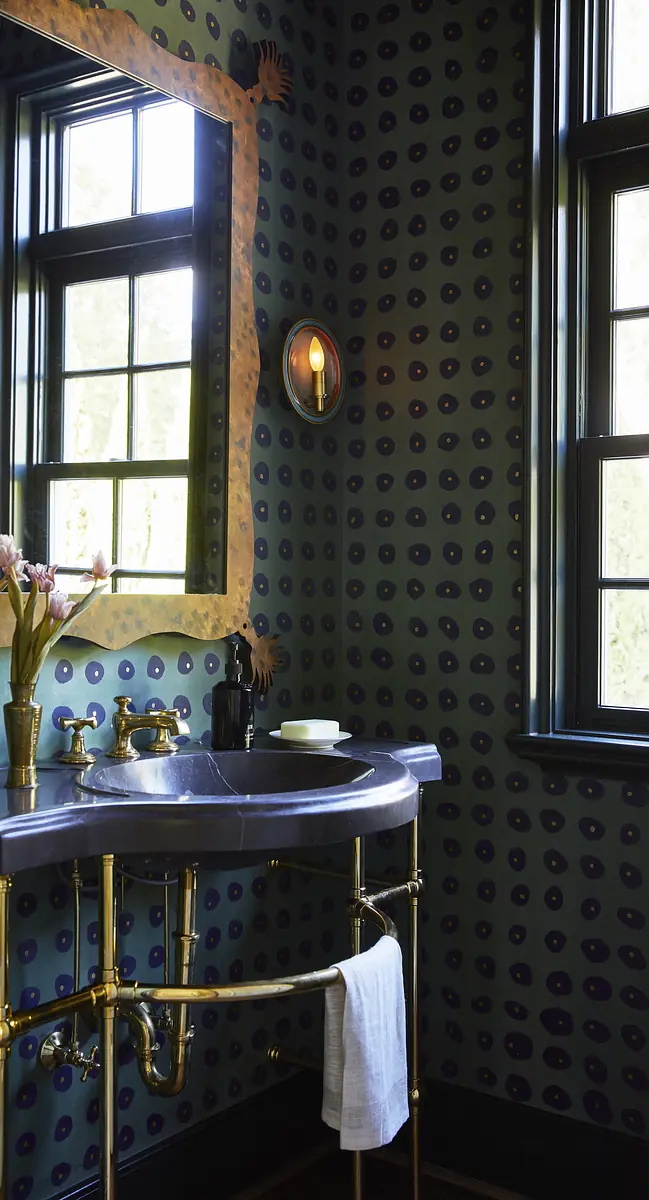 Bathroom with marble sink on brass frame, dark patterned wallpaper, two windows, and flowers in a vase.