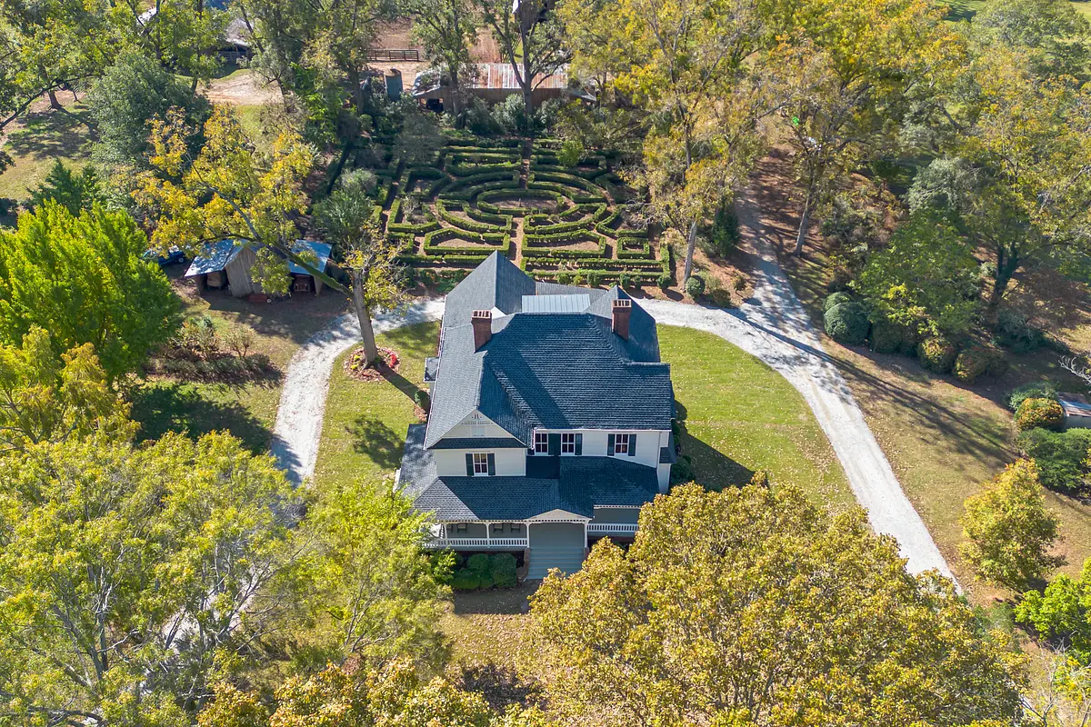 Aerial view of a house with gabled roof, grass yard, gravel driveway, and garden hedges in the background.