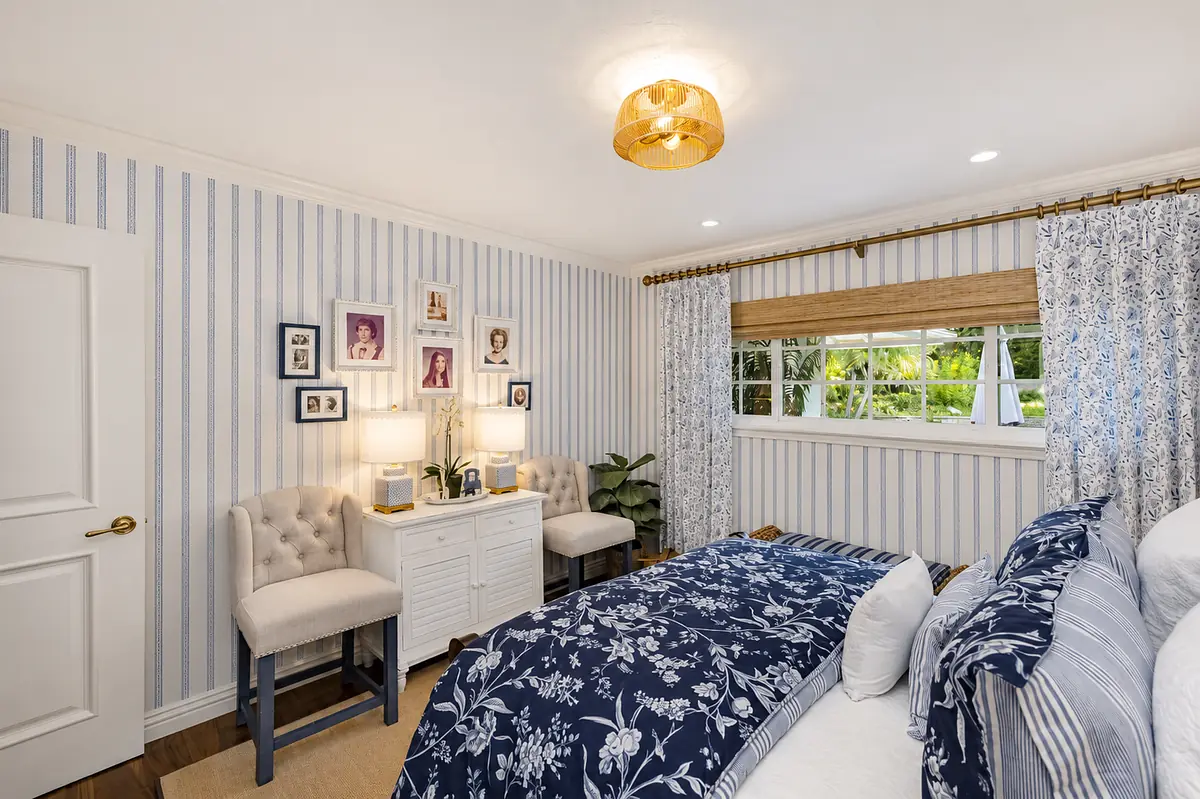 Bedroom with blue floral comforter, nightstands, cushioned chairs, striped wallpaper, and large window.