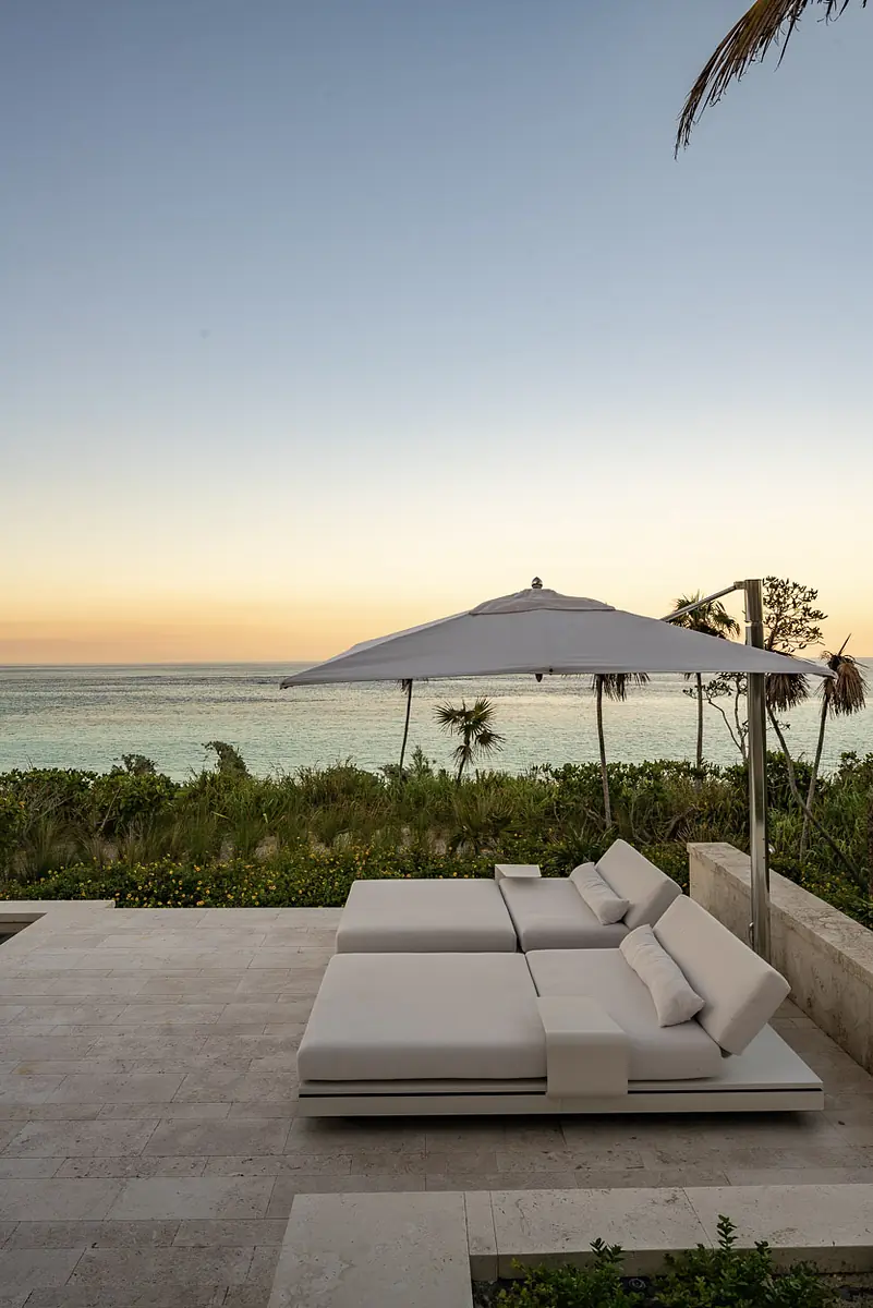 Patio with two white chaise lounges and large umbrella overlooking the ocean and sandy shore.