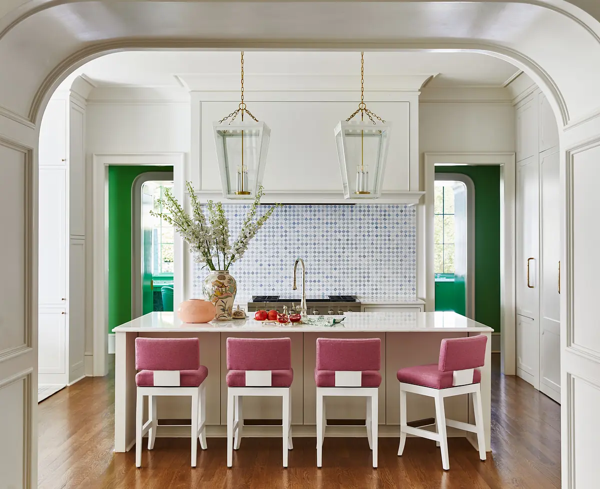 Kitchen with pink stools, white countertop, mosaic backsplash, green walls, and large windows providing natural light.