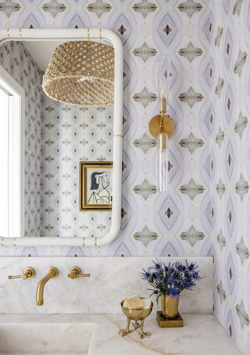 Bathroom with mirrored vanity, marble countertop, gold faucets, patterned wallpaper, and straw light fixture.