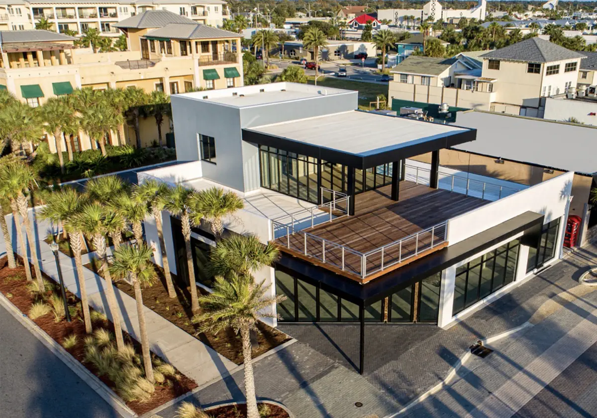 Modern building exterior with flat roof, large glass windows, wooden deck, and palm trees.