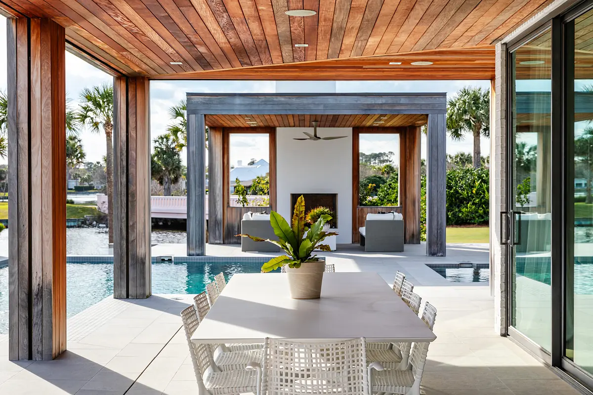 Patio with large table, white chairs, potted plants, swimming pool, and open wooden beams surrounded by greenery