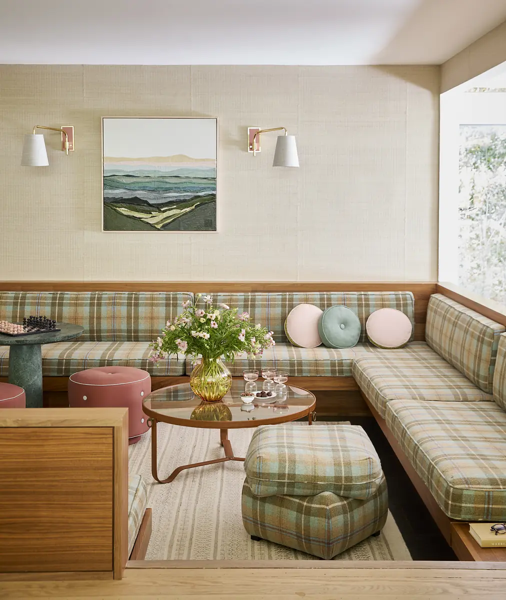 Living room with plaid sectional sofa, coffee table, cushions, flower vase, pink stools, wall sconces, and artwork.