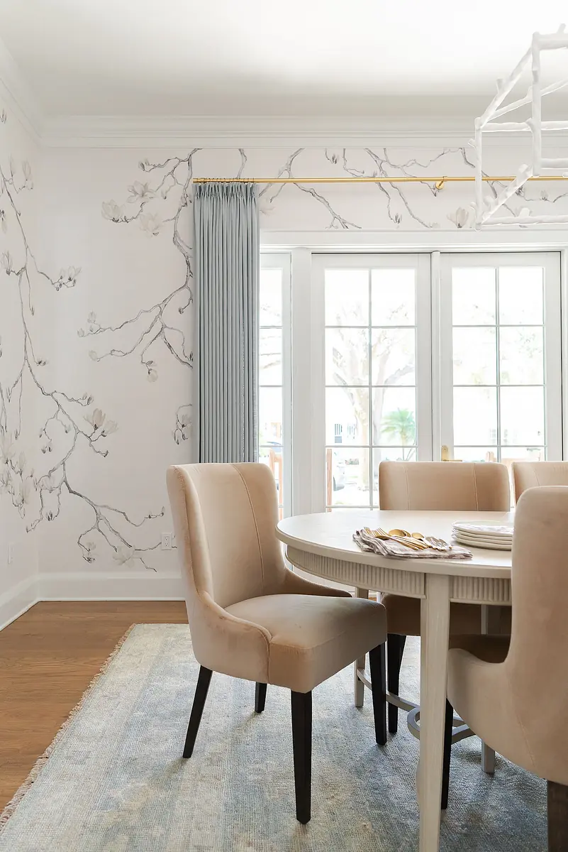 Dining room with round table, upholstered chairs, floral wallpaper, and large windows with sheer drapes.