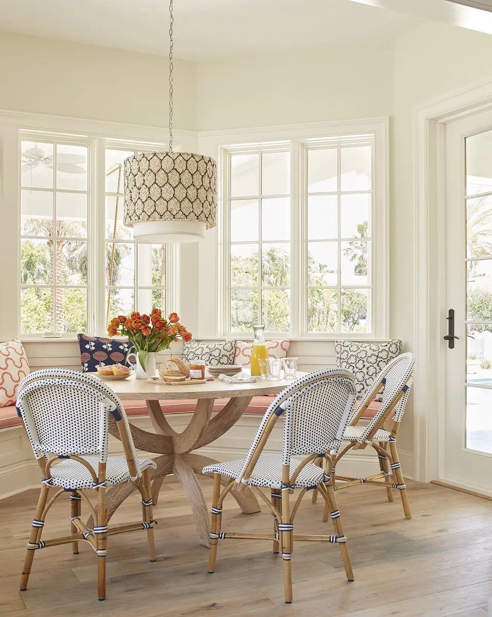 Dining room with round wooden table, rattan chairs, banquette, large windows, and a circular pendant light