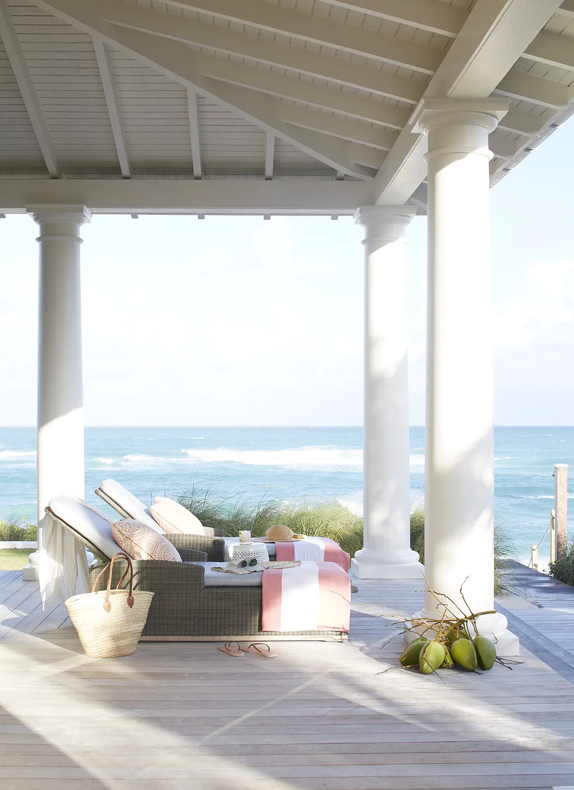 Patio with wicker lounge chair, side table, woven basket, ocean view, and white columns.