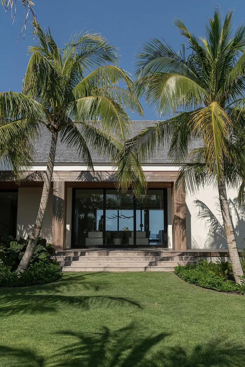 Exterior front view of a house with glass door, wooden framing, palm trees, and a green lawn.