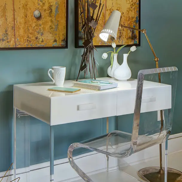 Office with white desk, clear acrylic chair, framed artworks, mug, notepad, and green plant on blue wall.